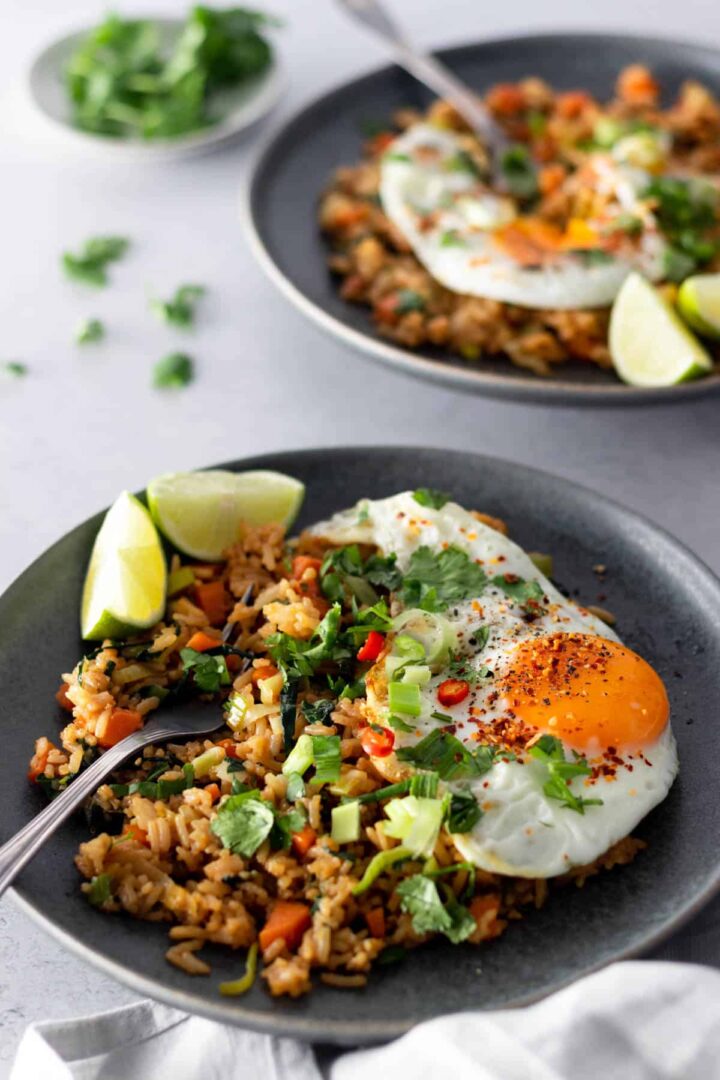 a grey plate with rice, cubed carrots, chopped green onions, leek, chopped cabbage, a fried egg with pepper, chili, chopped coriander, lime wedges and a fork on the side. Another plate out of focus. A small plate with chopped coriander on the side