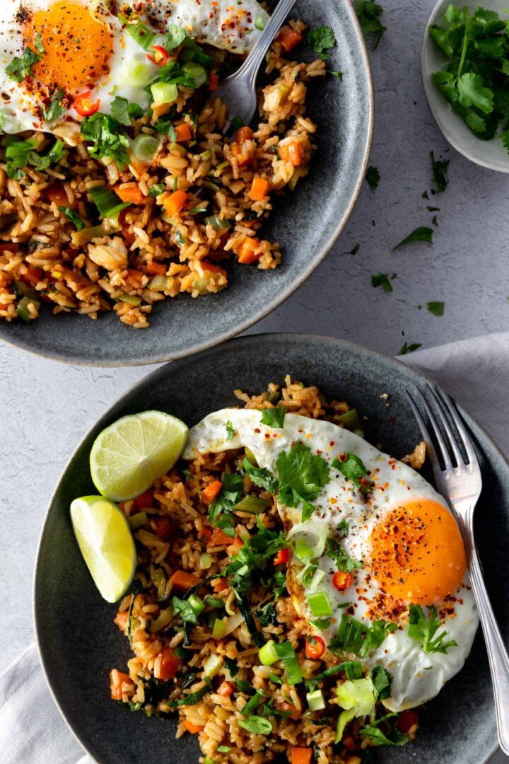 two grey plates with Nasi Goreng rice, cubed carrots, chopped green onions, leek, chopped cabbage, a fried egg with pepper, chili, chopped coriander, lime wedges, a fork on the side and a small plate with chopped coriander on the right top corner