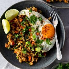 two grey plates with Nasi Goreng rice, cubed carrots, chopped green onions, leek, chopped cabbage, a fried egg with pepper, chili, chopped coriander, lime wedges, a fork on the side and a small plate with chopped coriander on the bottom right corner next to a grey kitchen towel