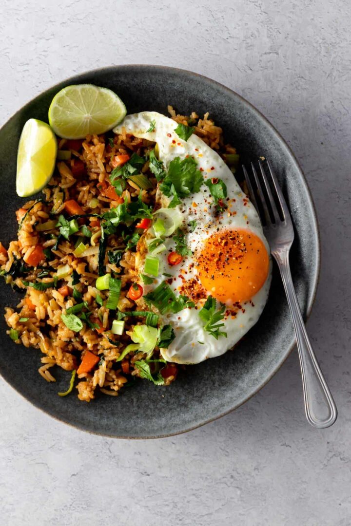 a grey plate with rice, cubed carrots, chopped green onions, leek, chopped cabbage, a fried egg with pepper, chili, chopped coriander, lime wedges and a fork on the side