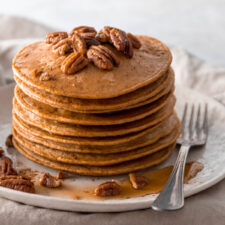 a plate with a stack of pancakes with pecans and maple syrup on top, a fork on the side and on top of a beige kitchen towel