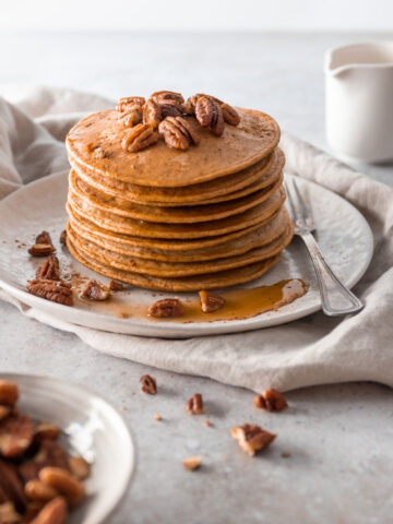 a plate with a stack of pancakes with pecans and maple syrup on top, a fork on the side and on top of a beige kitchen towel. A jug out of focus and a small bowl with pecans out of focus