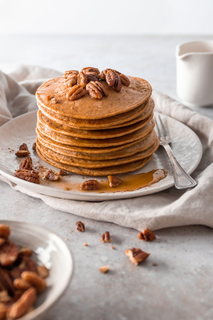 a plate with a stack of pancakes with pecans and maple syrup on top, a fork on the side and on top of a beige kitchen towel. A jug out of focus and a small bowl with pecans out of focus
