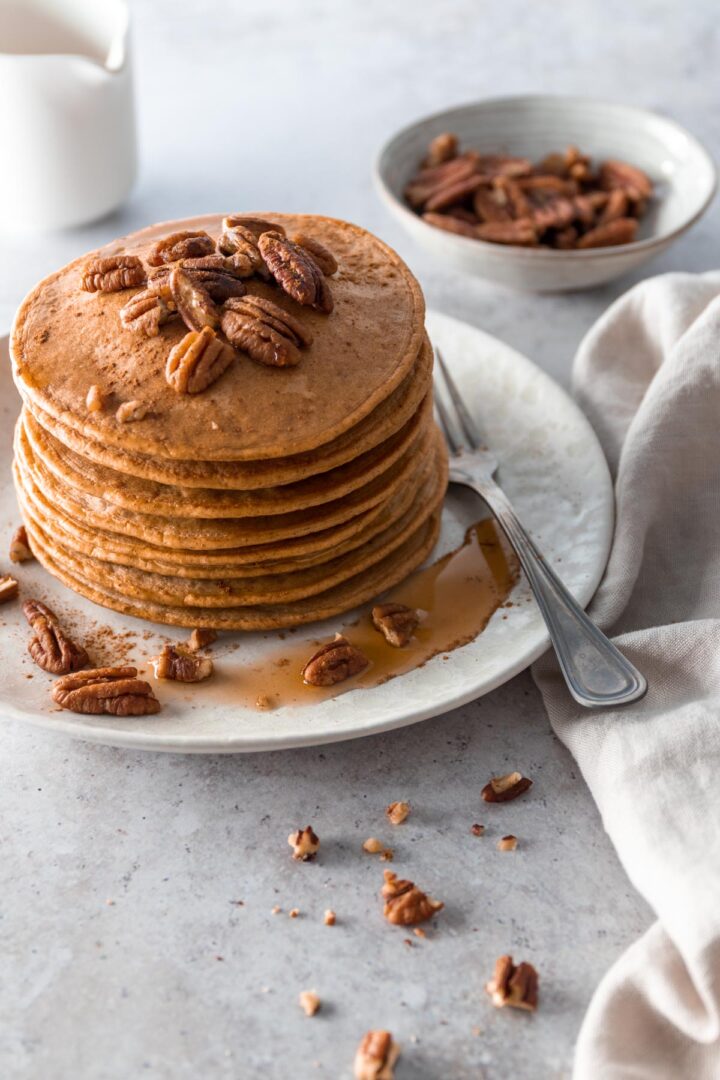 a plate with a stack of pancakes with pecans and maple syrup on top, a fork on the side and on top of a beige kitchen towel. A jug out of focus and a small bowl with pecans out of focus