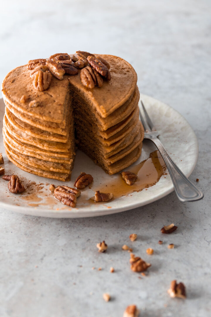 a plate with a stack of pancakes, sliced, with pecans and maple syrup on top, a fork on the side