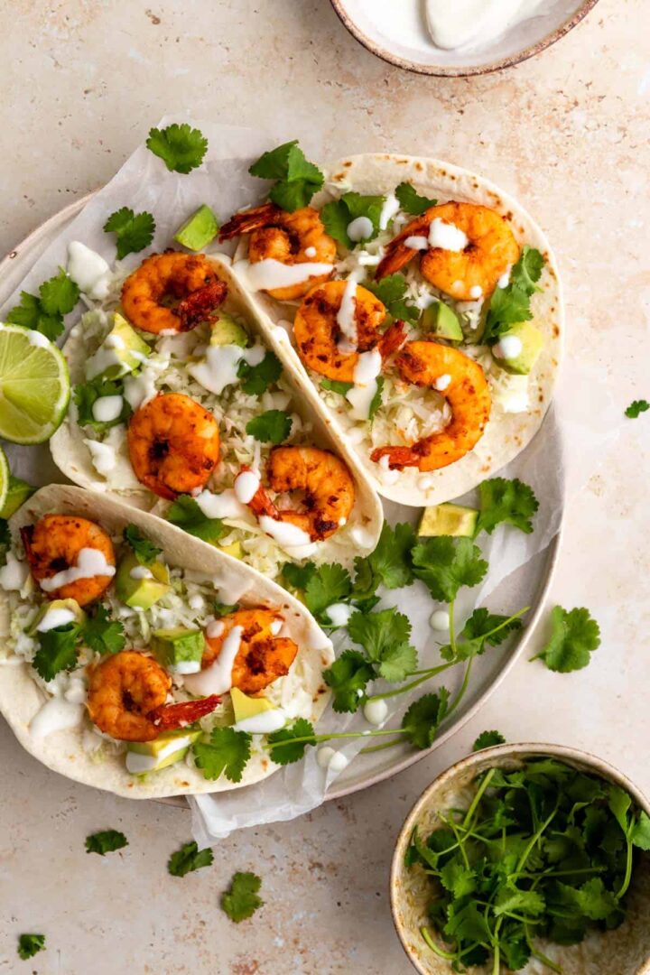 three shrimp tacos served on a plate and garnished with coriander and cream and a small bowl with more coriander leaves
