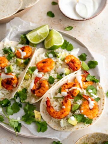 three shrimp tacos served on a plate and garnished with coriander and cream and two lime wedges on the side