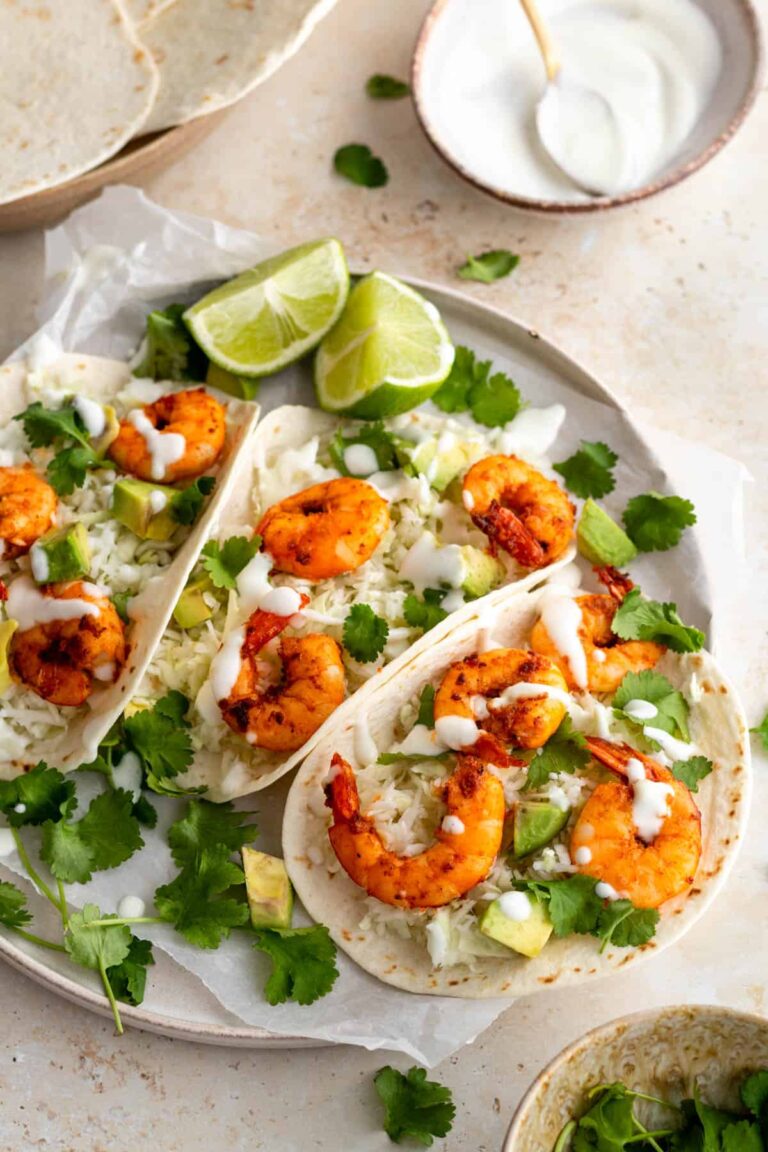 three shrimp tacos served on a plate and garnished with coriander and cream and two lime wedges on the side