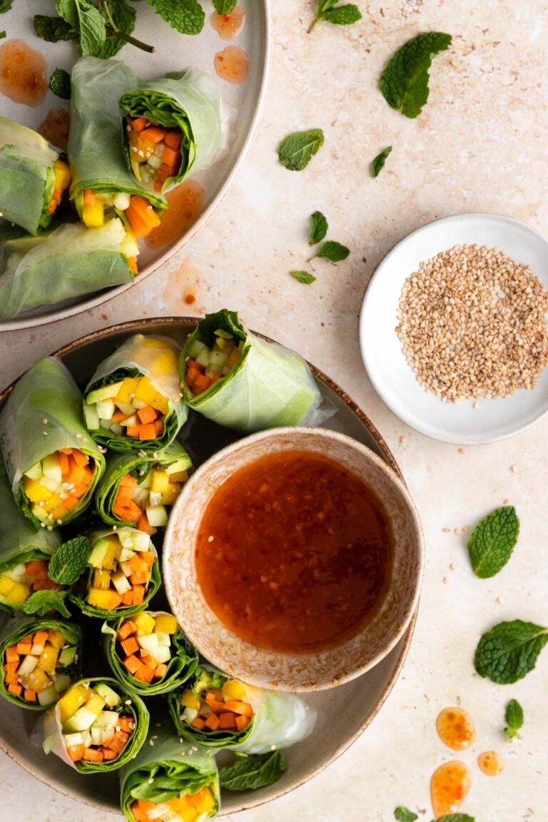 two plates with rice paper rolls with carrot, avocado, mango, cucumber, lettuce and mint. A bowl with sweet chili sauce and a small plate with sesame seeds