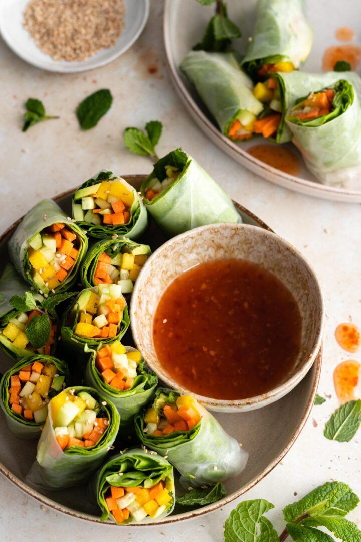 two plates with rice paper rolls with carrot, avocado, mango, cucumber, lettuce and mint. A bowl with sweet chili sauce and a small plate with sesame seeds