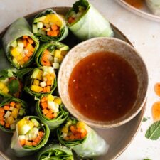 two plates with rice paper rolls with carrot, avocado, mango, cucumber, lettuce and mint. A bowl with sweet chili sauce and more mint on top of a board