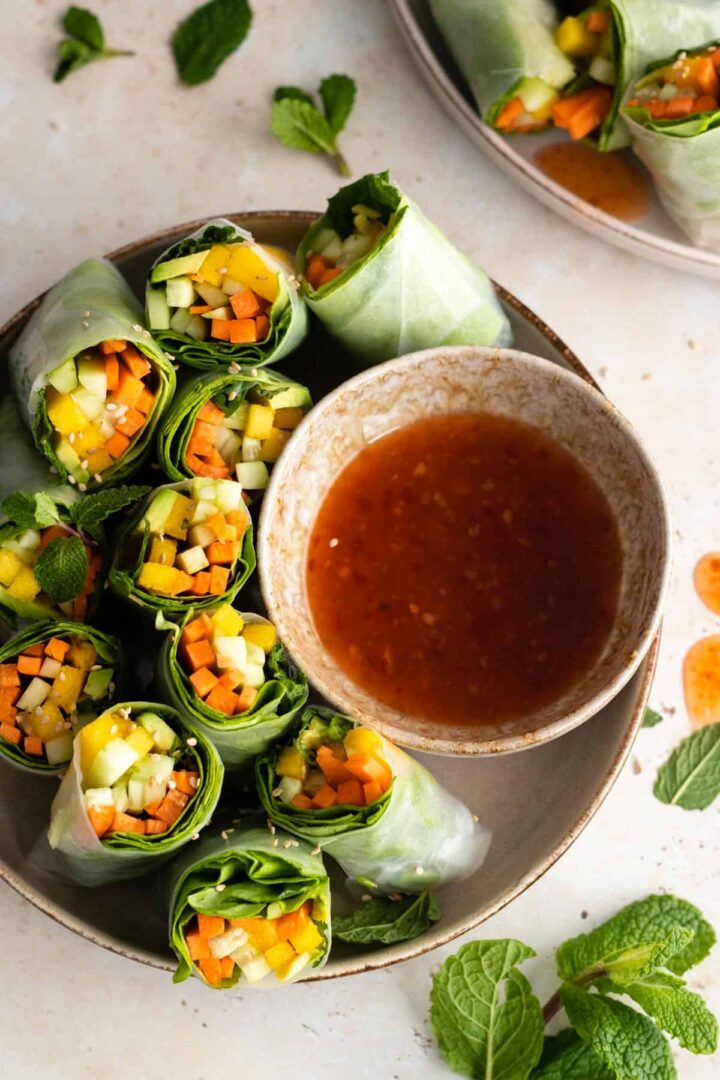 two plates with rice paper rolls with carrot, avocado, mango, cucumber, lettuce and mint. A bowl with sweet chili sauce and more mint on top of a board