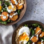 two sweet potato buddha bowls with baked sweet potato, mixed greens, quinoa, avocado, poached egg, white dressing and black sesame seeds on top