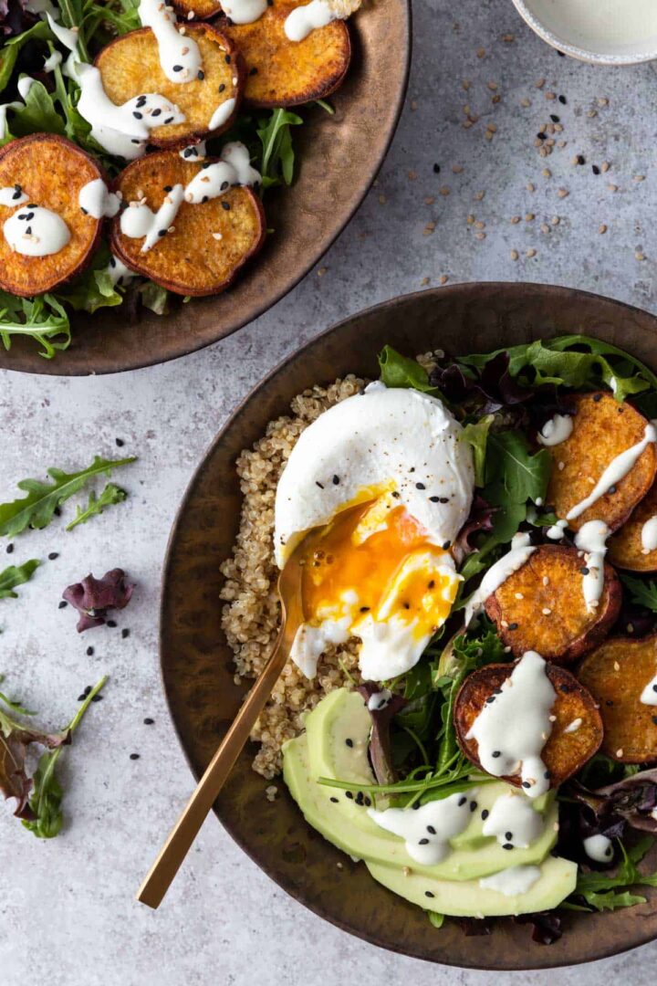 two bowls with mixed greens, quinoa, sliced avocado, sweet potato, poach egg and dressing on top and a small bowl with dressing on the corner