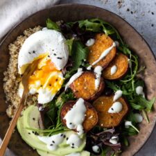 close up sweet potato buddha bowl with quinoa, avocado, sweet potato, mixed greens, white dressing and sesame seeds on top