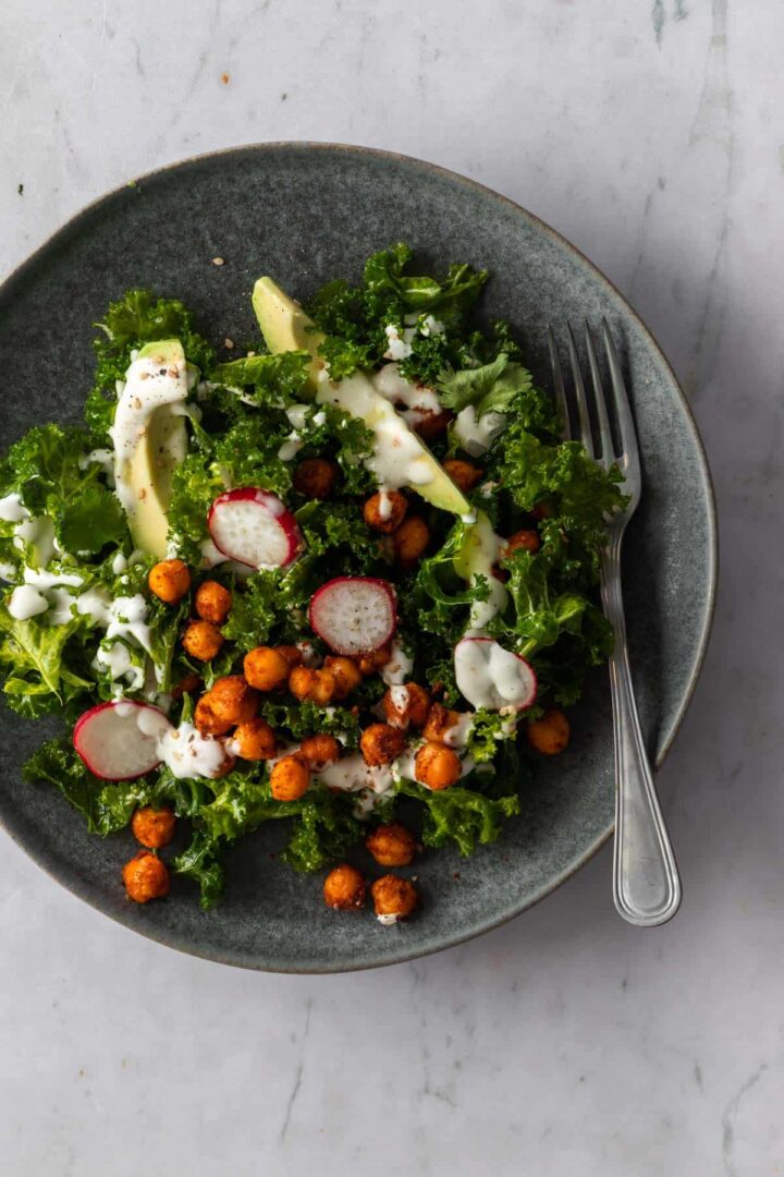 massaged kale, sliced avocado, radishes, chickpeas on a plate with dressing on top and a fork on the side