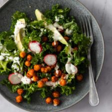 massaged kale, sliced avocado, radishes, chickpeas on a plate with dressing on top and a fork on the side
