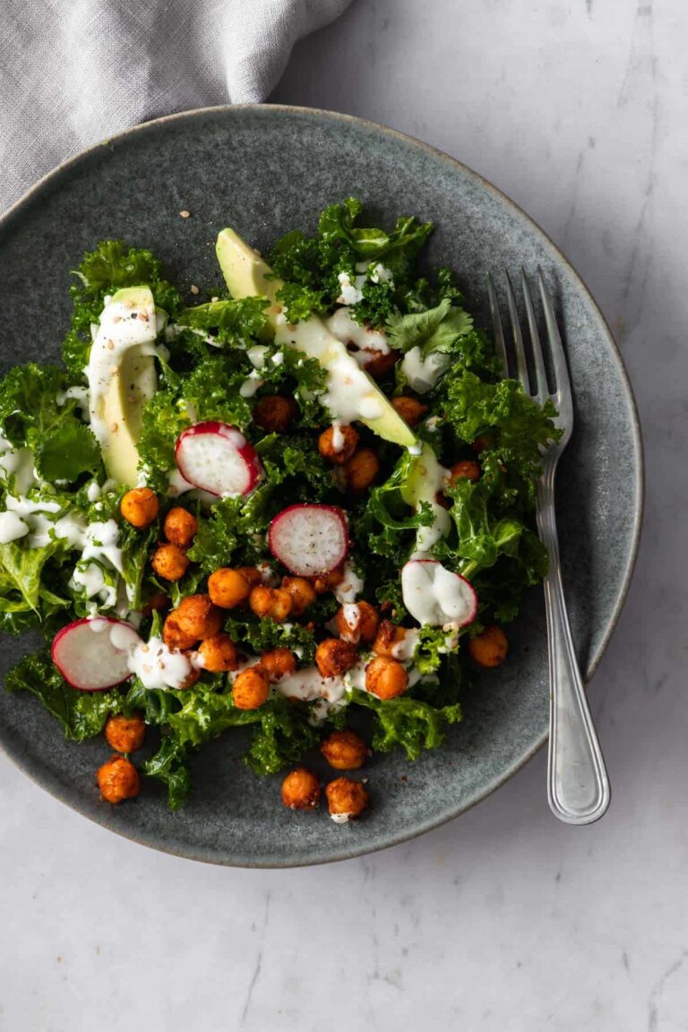 massaged kale, sliced avocado, radishes, chickpeas on a plate with dressing on top and a fork on the side