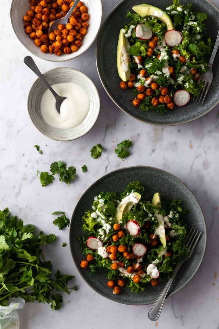 two plates with massaged kale salad, a bowl with dressing, a bowl with chickpeas and coriander on top of a marble table