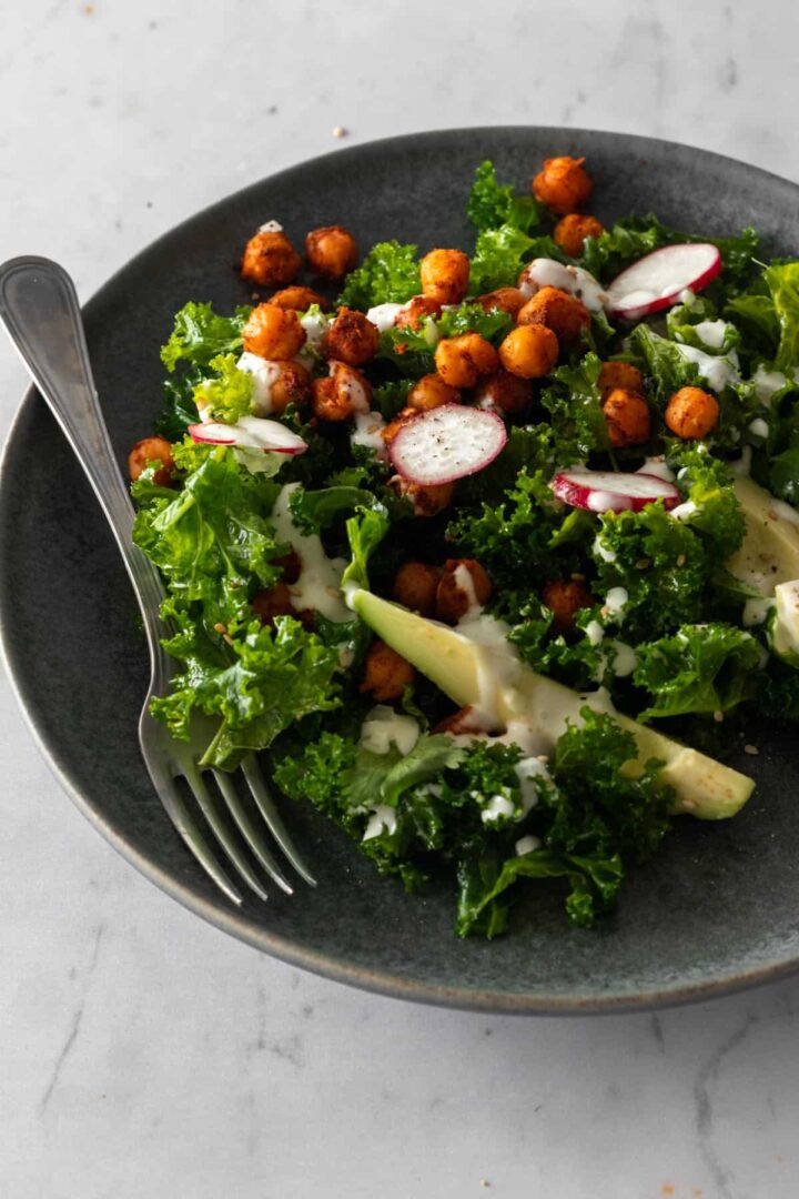 massaged kale, sliced avocado, radishes, chickpeas on a plate with dressing on top and a fork on the side