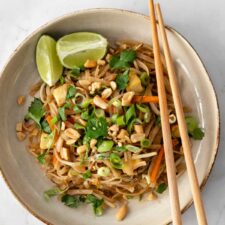 a plate with noodles, julienned carrots, chopped green onions, been sprouts, cubed tofu, onion, chopped coriander, peanuts, lime and chopsticks on the side