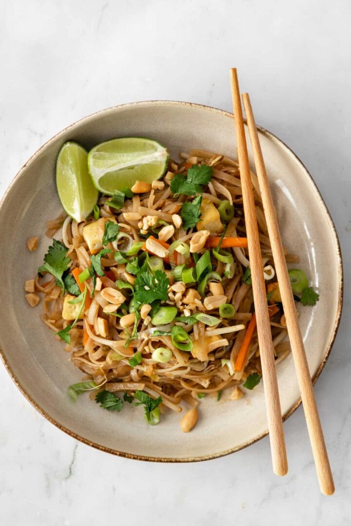 a plate with noodles, julienned carrots, chopped green onions, been sprouts, cubed tofu, onion, chopped coriander, peanuts, lime and chopsticks on the side