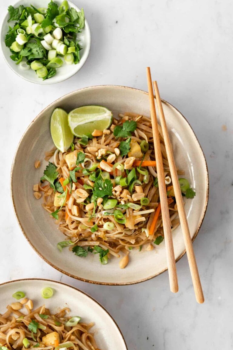 a plate with noodles, julienned carrots, chopped green onions, been sprouts, cubed tofu, onion, chopped coriander, peanuts, lime and chopsticks on the side. Another plate on the bottom left corner and a small plate with chopped green onions and coriander on the top left corner