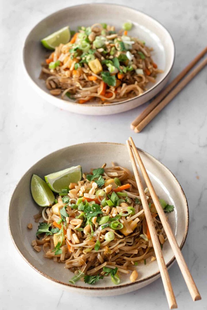 two plates with noodles, julienned carrots, chopped green onions, been sprouts, cubed tofu, onion, chopped coriander, peanuts, lime and chopsticks on the side