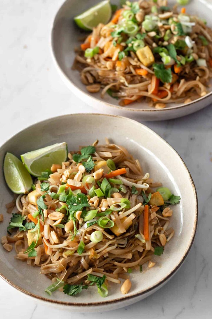 two plates with pad Thai noodles, julienned carrots, chopped green onions, been sprouts, cubed tofu, onion, chopped coriander, peanuts, lime and chopsticks on the side