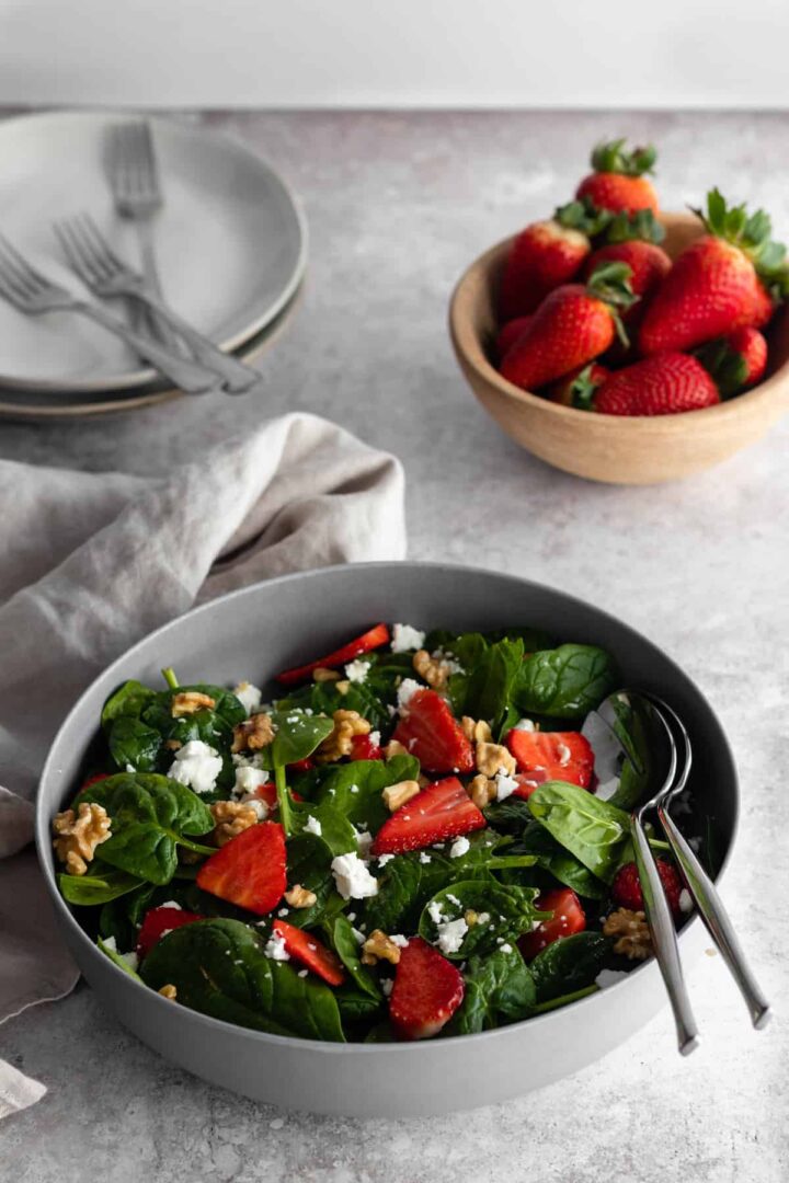 a grey salad bowl with spinach, strawberries, feta cheese and walnuts. A small bowl with more strawberries, two plates and three forks and a kitchen towel on top of a backdrop