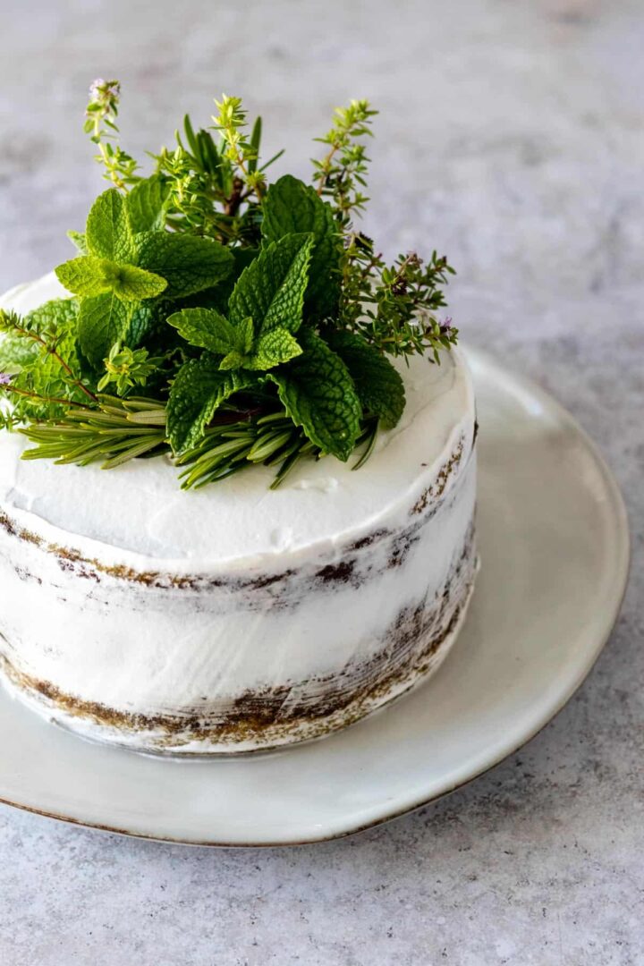 a plate with a semi naked cake covered with a whipped cream frosting and decorated with fresh herbs on top