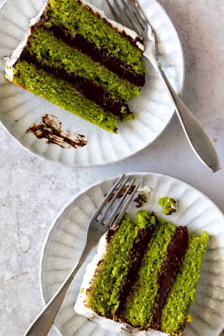 2 plates with two slices of a three layered green watercress cake with chocolate ganache and whipped cream frosting and two forks on the side