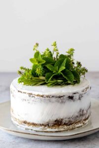 a plate with a semi naked cake covered with a whipped cream frosting and decorated with fresh herbs on top