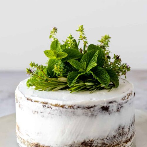 a plate with a semi naked cake covered with a whipped cream frosting and decorated with fresh herbs on top