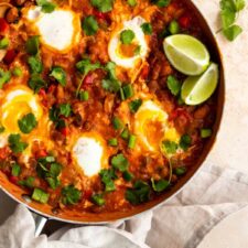 A pan with eggs in tomato sauce, bell peppers, coriander and lime wedges