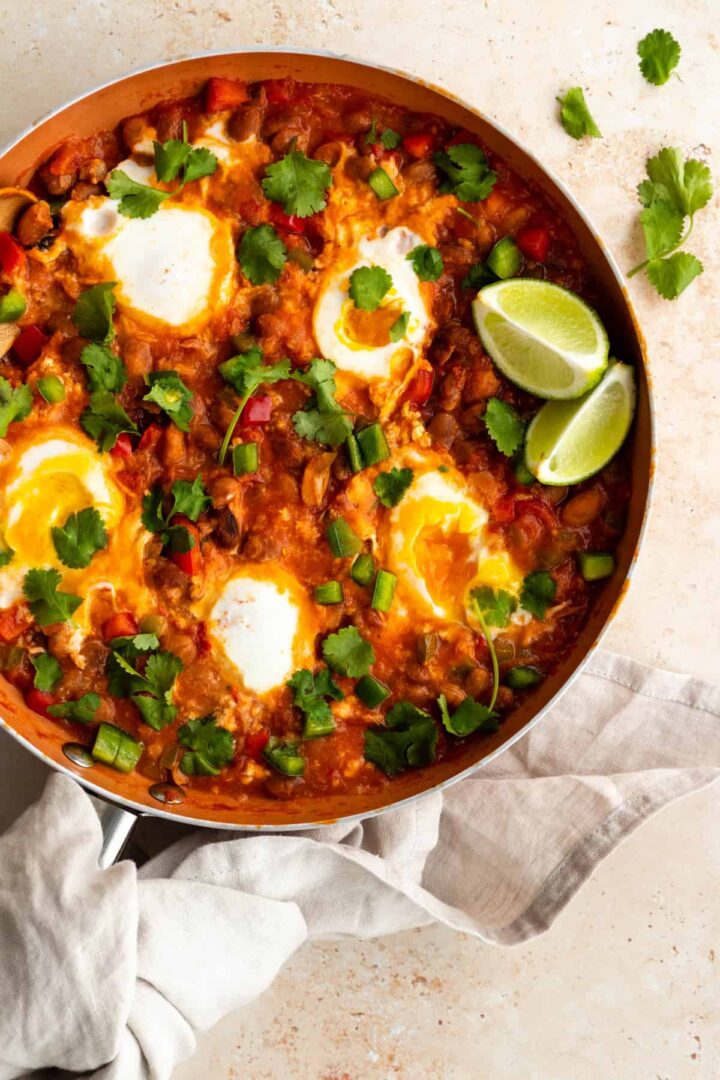 A pan with huevos rancheros in tomato sauce, bell peppers, coriander and lime wedges