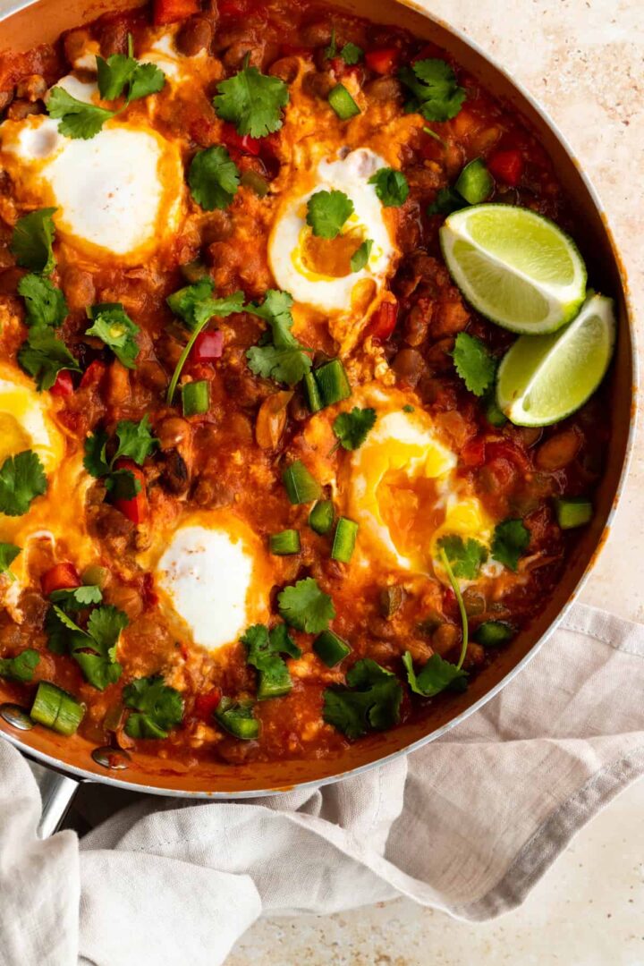 A pan with eggs in tomato sauce, bell peppers, coriander and lime wedges