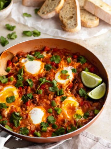 a pan with eggs, beans, tomatoes and bell pepper, garnished with coriander and lime wedges. Toasted bread on the top right corner and a bowl with more coriander on the top left corner