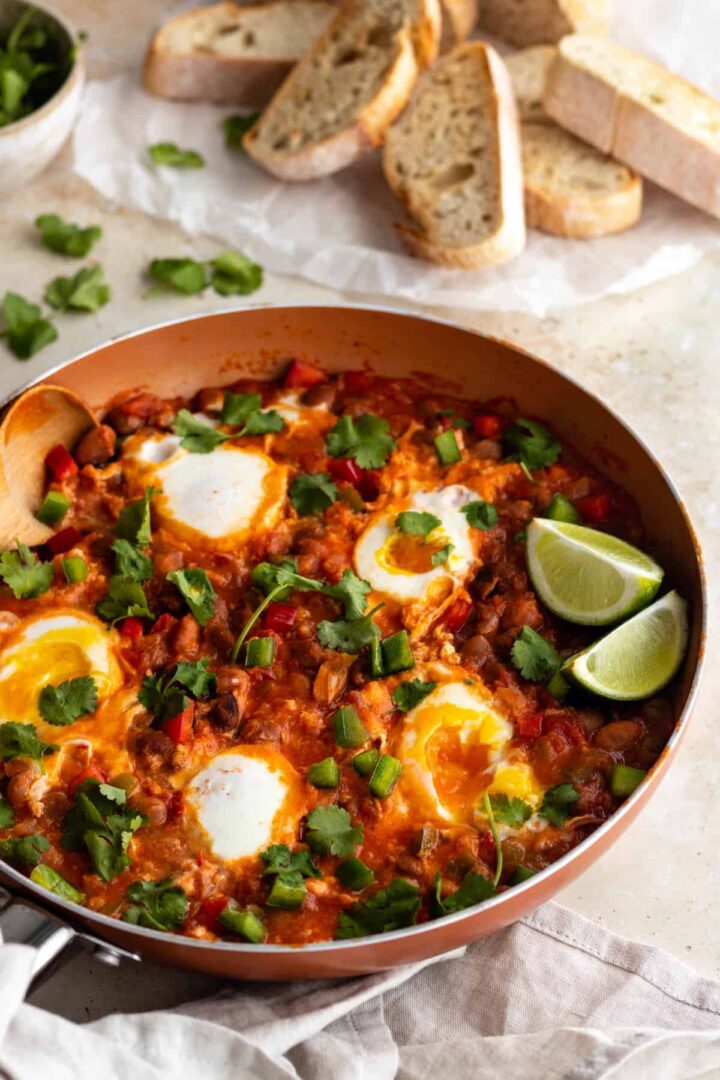 a pan with eggs, beans, tomatoes and bell pepper, garnished with coriander and lime wedges. Toasted bread on the top right corner and a bowl with more coriander on the top left corner