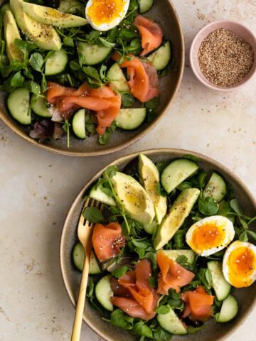 two bowls with mixed greens salad with smoked salmon, cucumber, avocado and boiled egg, a small bowl on the side with sesame seeds