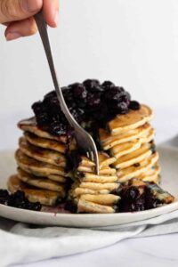 a plate with a stack of pancakes with blueberries and maple syrup on top and someone taking some with a fork