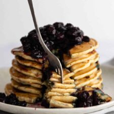 a plate with a stack of pancakes with blueberries and maple syrup on top and someone taking some with a fork