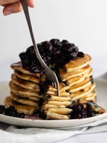 a plate with a stack of pancakes with blueberries and maple syrup on top and someone taking some with a fork