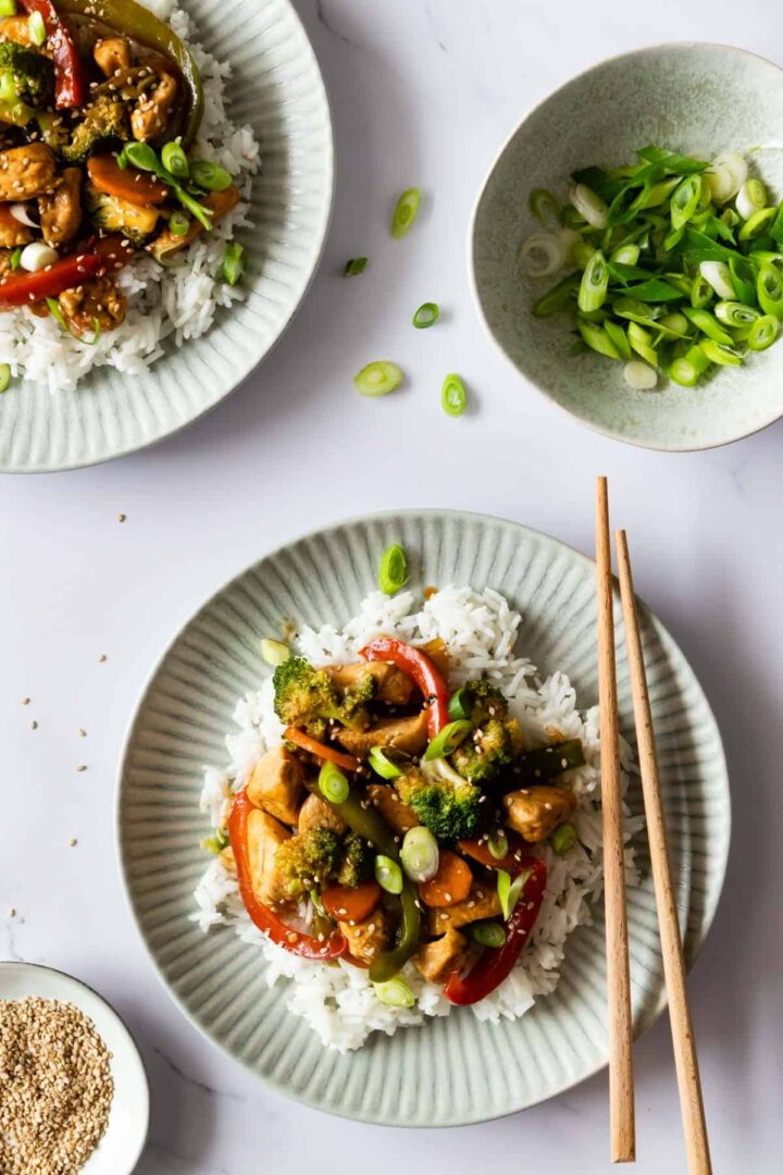 two plates served with rice, chicken, vegetables and garnished with sesame seeds as green onions, two small bowls on the side, one with sesame seeds and the other one with chopped green onion