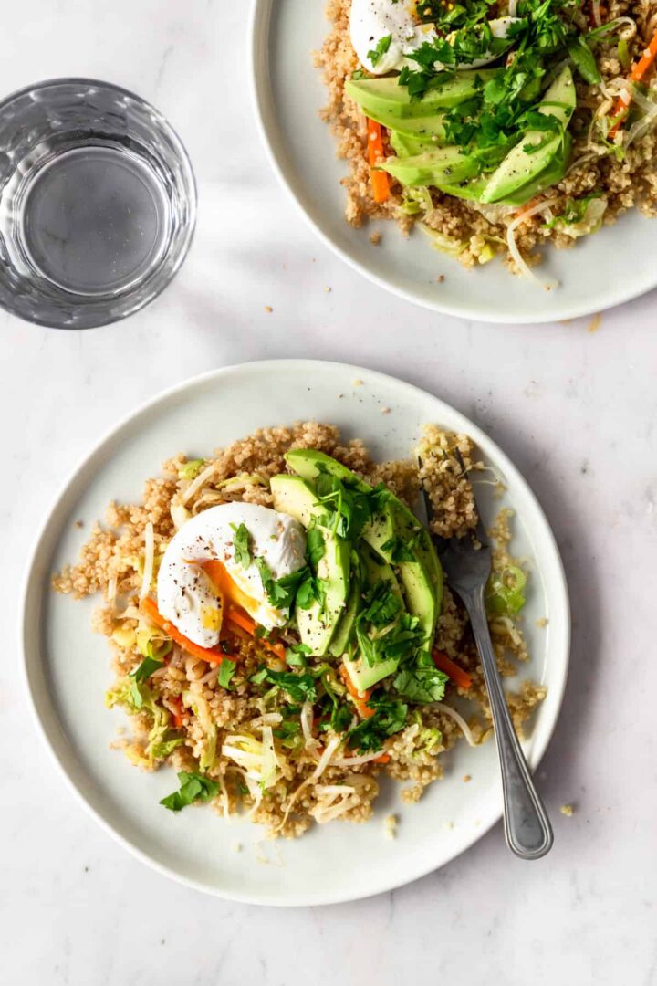 two plates served with quinoa stir fry with vegetables, poached egg, sliced avocado and chopped coriander on top. A glass of water on the side