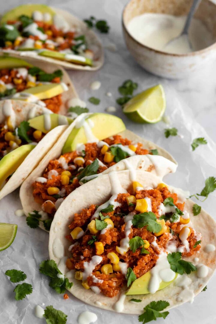 tortillas with quinoa, corn, coriander, avocado place on a parchment paper with lime wedges and topped with vegan dressing, a bowl with dressing on the top right corner