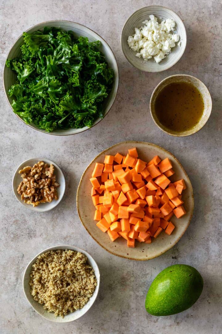 a bowl with shredded kale, a bowl with feta cheese, a bowl with dressing, a small plate with walnuts, a plate with cubbed sweet potato, a bowl with cooked quinoa and one avocado