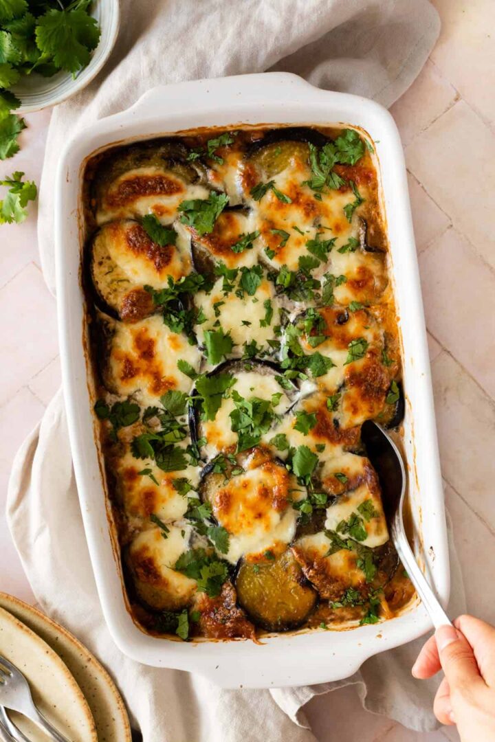 vegetable moussaka being served on a baking dish with chopped coriander on top, a bowl with more coriander on the side and forks on the bottom left corner