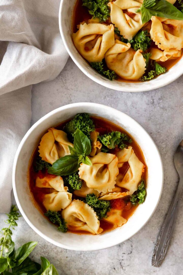 two bowls served with tortellini soup with kale, tomatoes and garnished with fresh basil