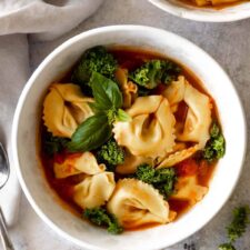 a bowl served with tortellini soup with kale, tomatoes and garnished with fresh basil. Another bowl on the top right corner with the same soup.
